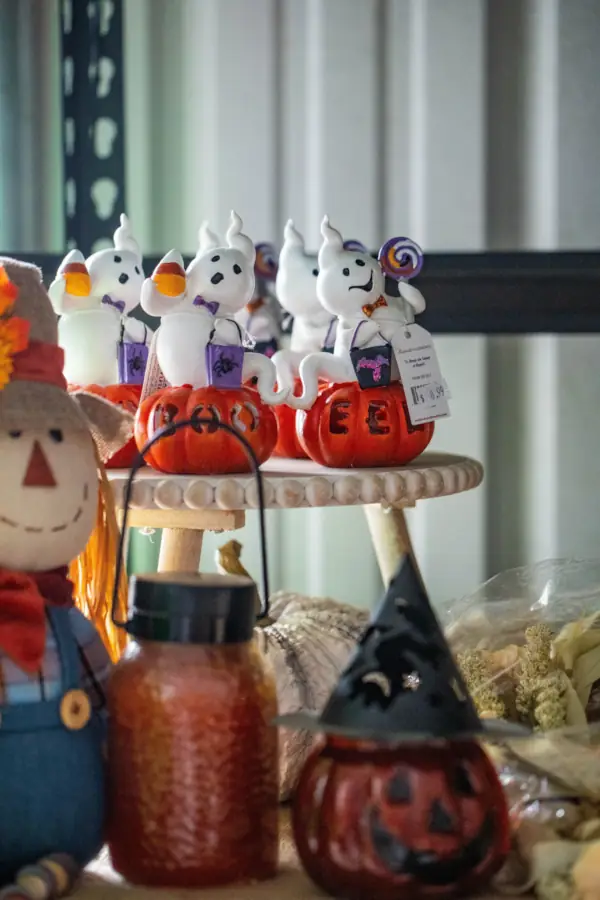 Halloween-themed display with small ghost figurines atop orange jack-o'-lantern bases, a scarecrow doll, and pumpkin-shaped jars. The mood is festive and whimsical.