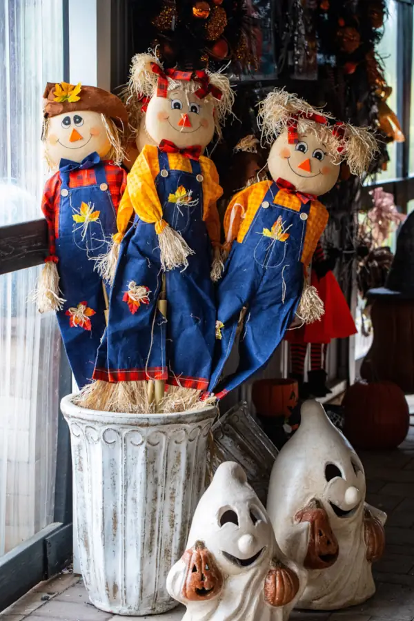 Three cheerful scarecrow decorations in denim overalls and straw hair sit in a pot beside two smiling ceramic ghosts holding pumpkins. Festive and playful Halloween vibe.