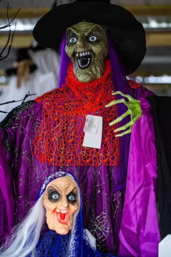 A green-faced witch decoration with a wide grin and purple hair wears a black hat and vibrant red and purple clothes. The small, eerie doll below shares a similar menacing expression. The scene evokes a spooky Halloween atmosphere.
